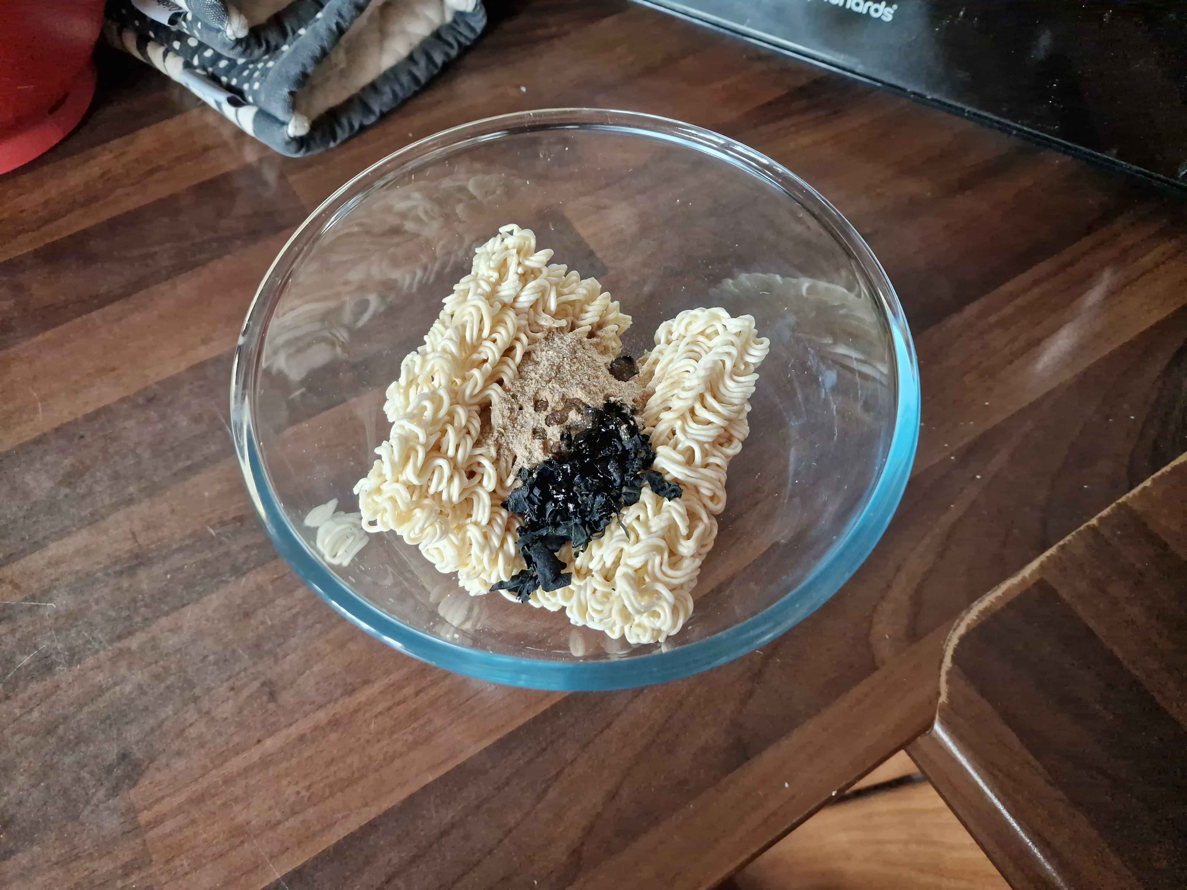 A glass bowl with the unprepared noodle block, powder, sesame oil and seaweed.