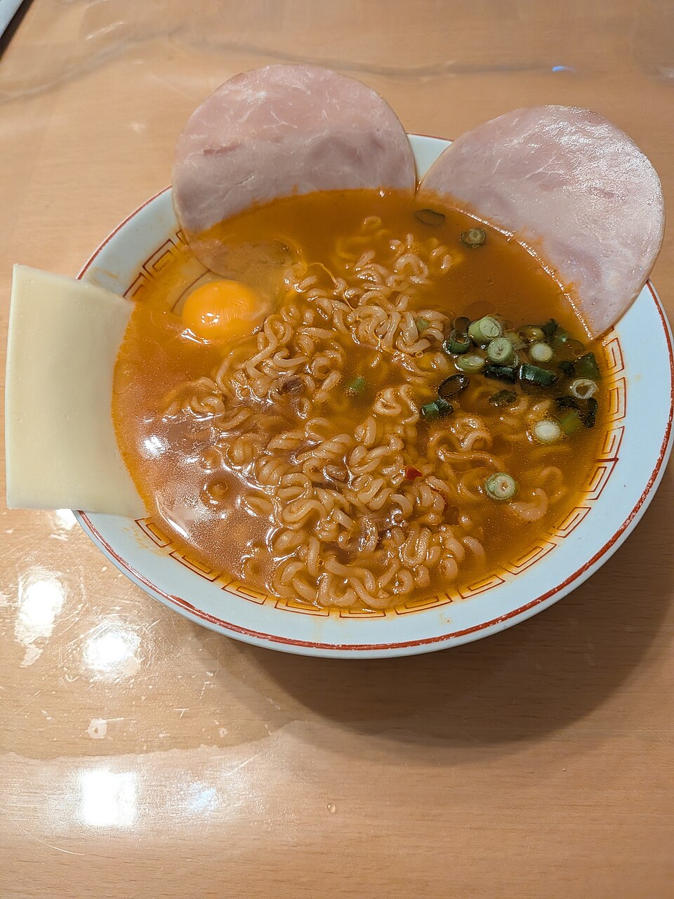 A bowl of Shin Ramyun noodles adorned with what appears to be slices deli ham and cheese around the sides.