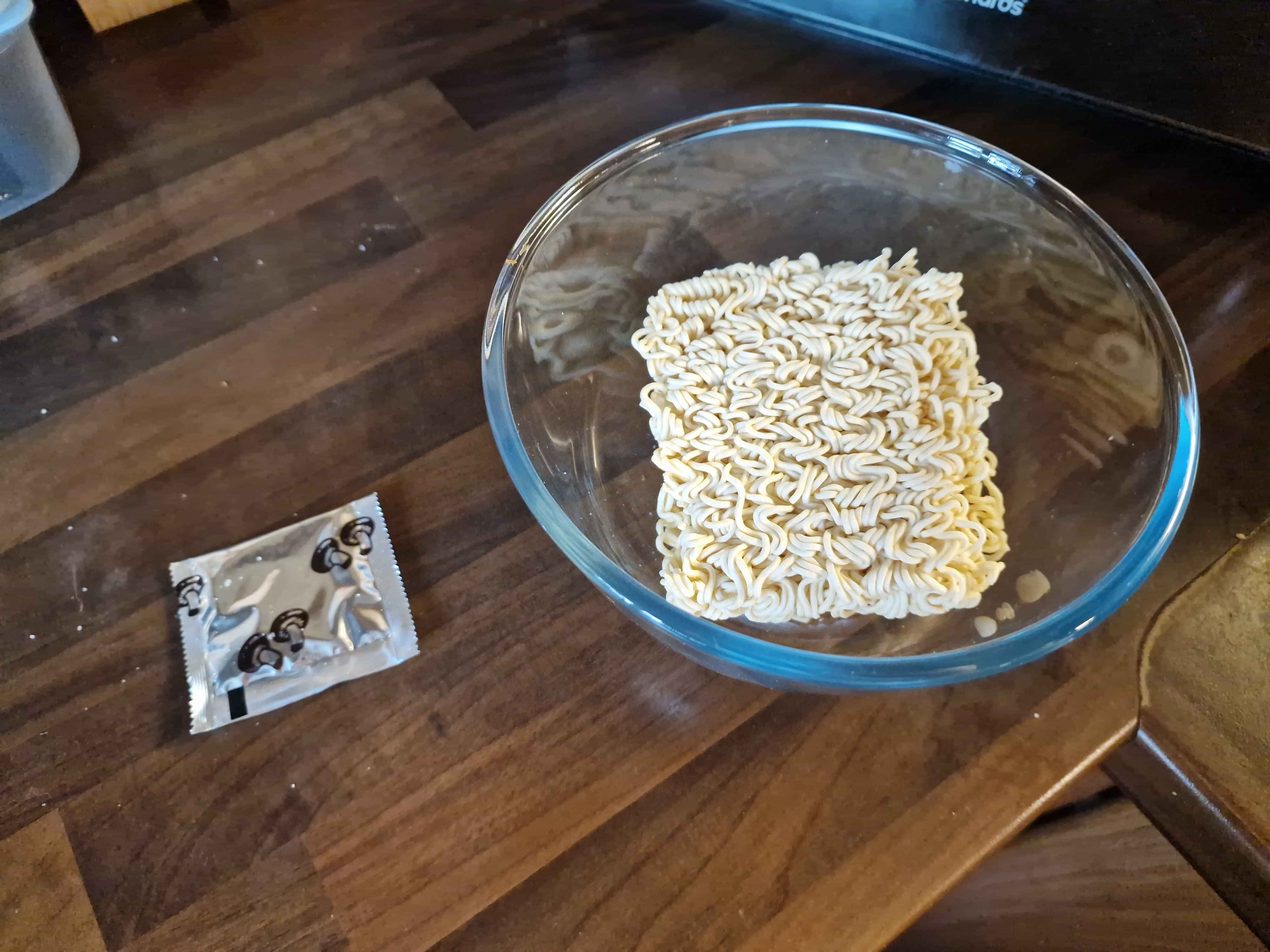 A glass bowl containing the raw noodle brick, next to the seasoning packet.