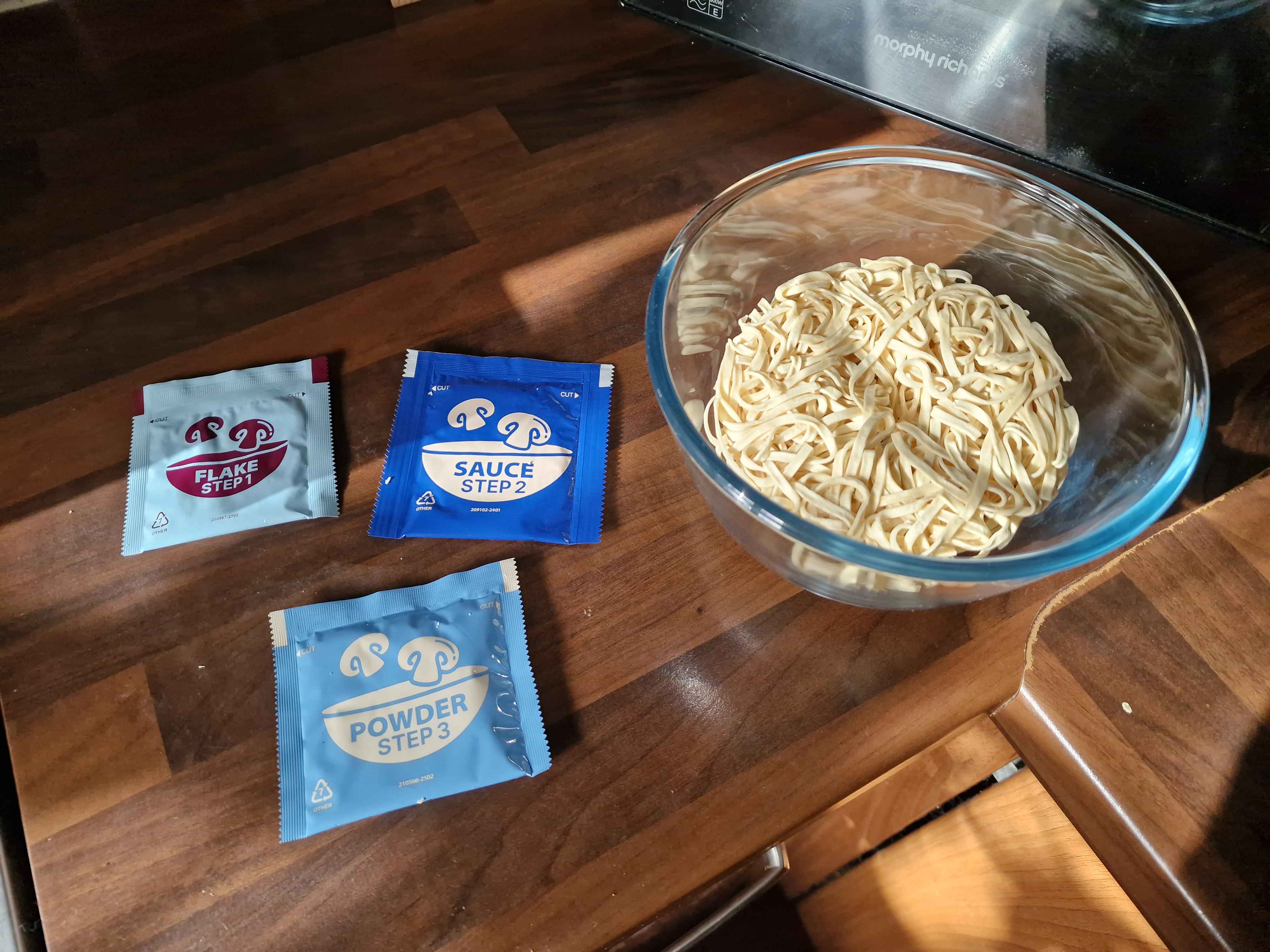 The dry, round flat noodle brick inside a glass bowl, next to three numbered sachets each for flakes, sauce and powder.