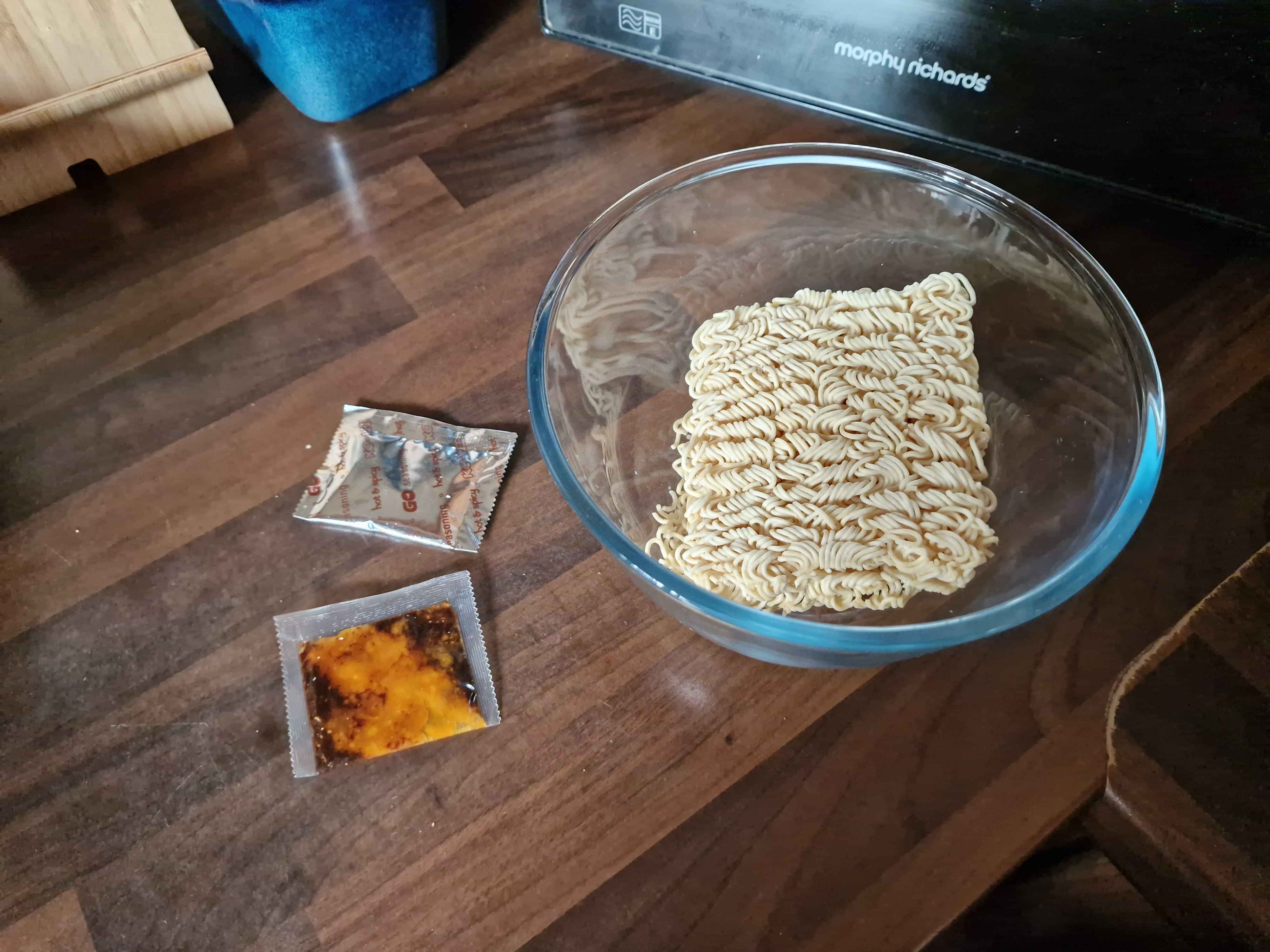 The contents of the ramen pack, a block of noodles in a glass bowl, a metallic sachet of powder and a clear sachet of oil with chili crisps.