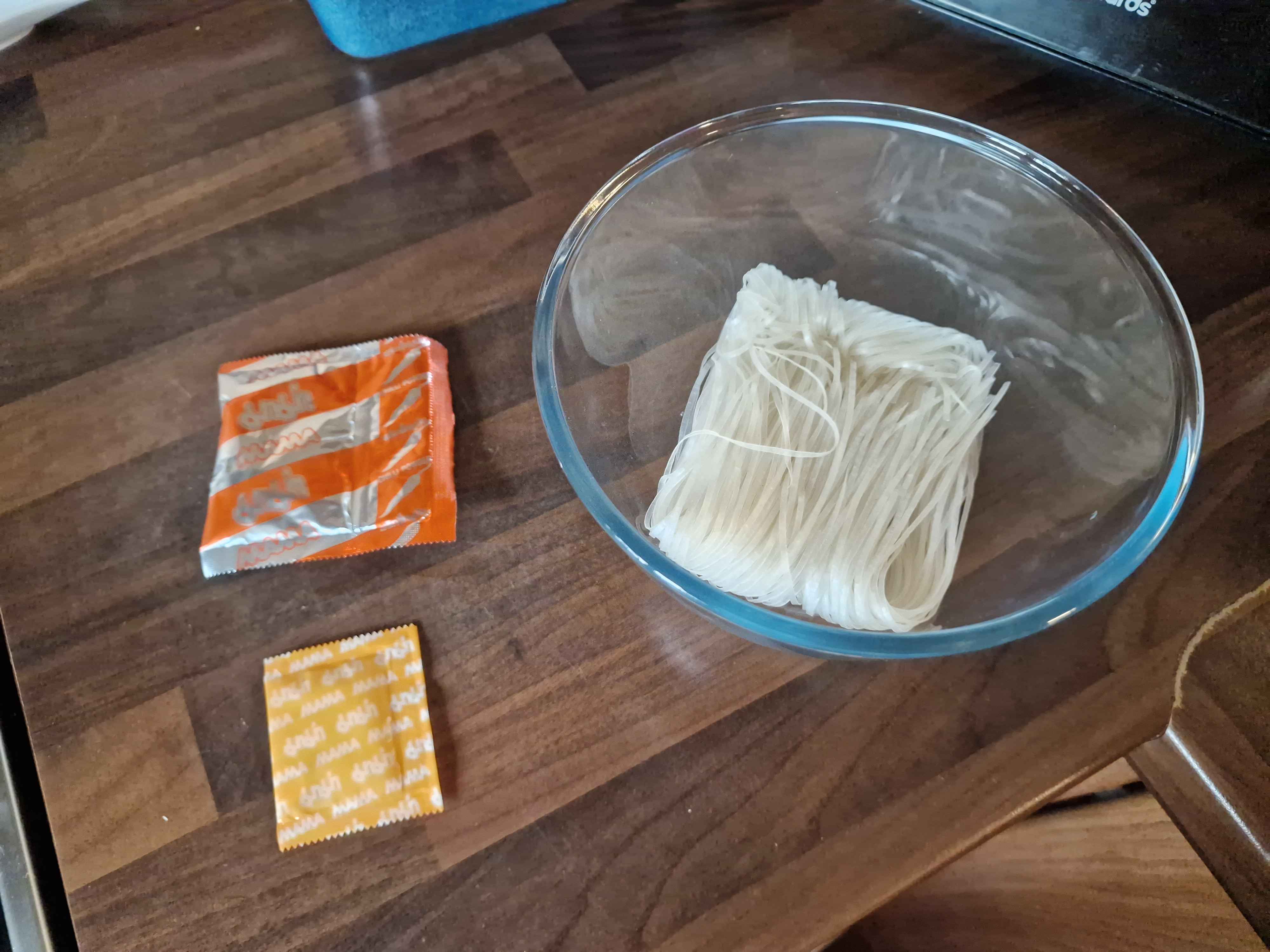 A glass bowl containing an uncooked rice noodle brick, next to two packets carrying the Mama logo.