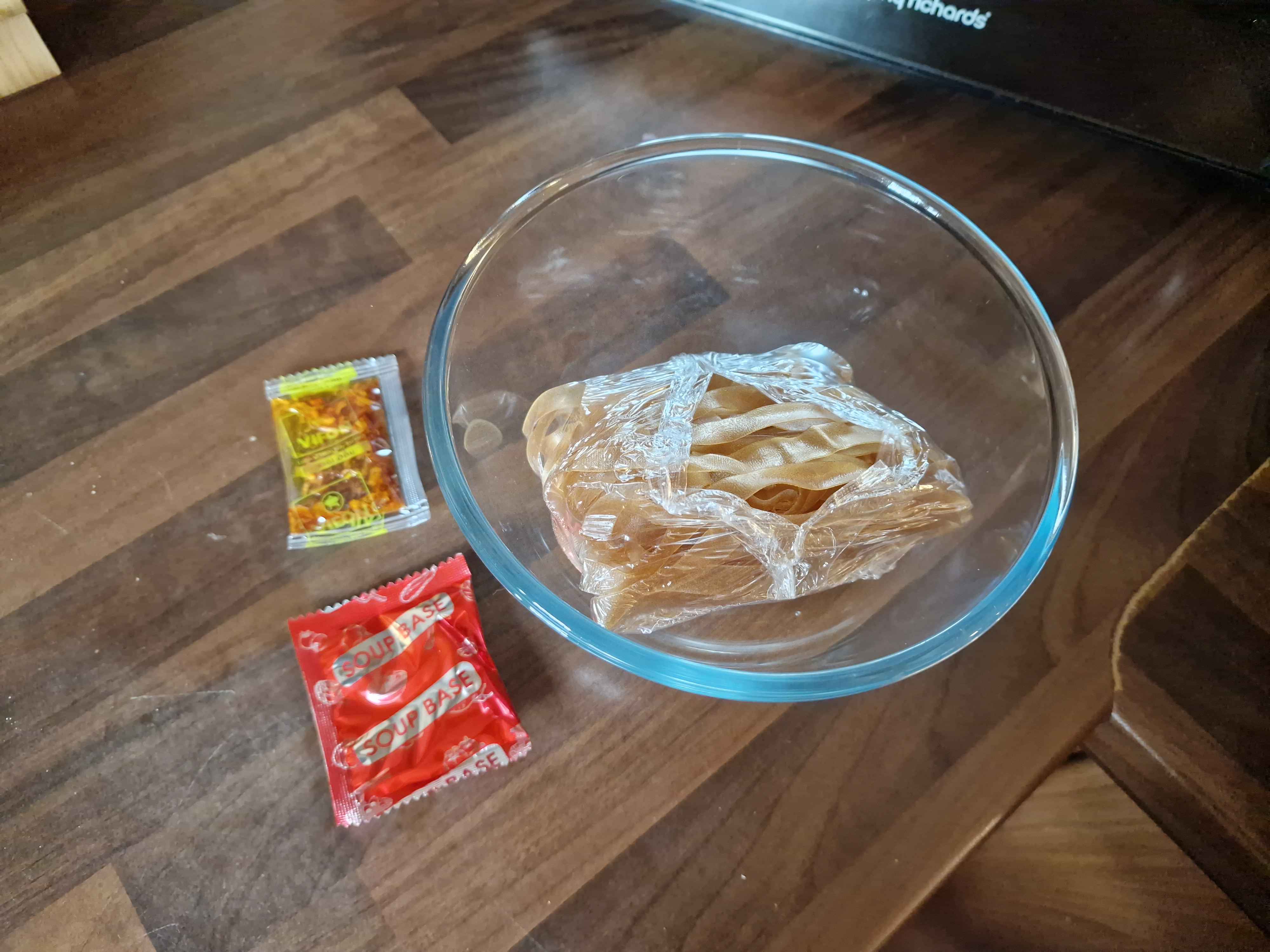 The contents of the noodle package, two sachets each with crispy oil and soup base, and the brown rice noodles wrapped in plastic inside the bowl.