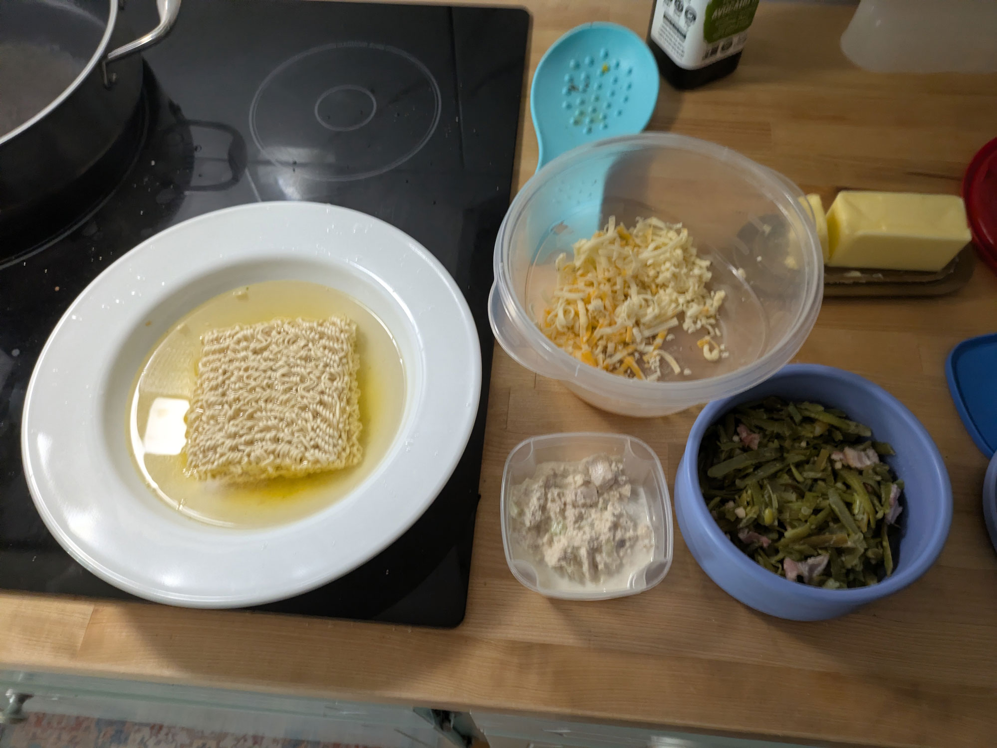 A bowl of noodles stewing in hot water, a container of cheese, a small container of tuna, and a medium bowl of green beans and bacon.