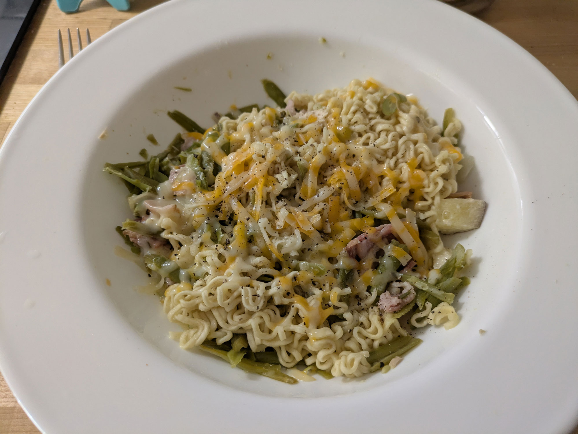 A large bowl containing ramen noodles, green beans, chopped bacon, tuna salad, and shredded cheese.